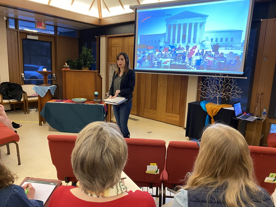 White woman with dark brown hair standing in front of a town hall meeting. The projector screen behind her shows a crowd in front of the Supreme Court of the United States and the League of Women Voters logo. 